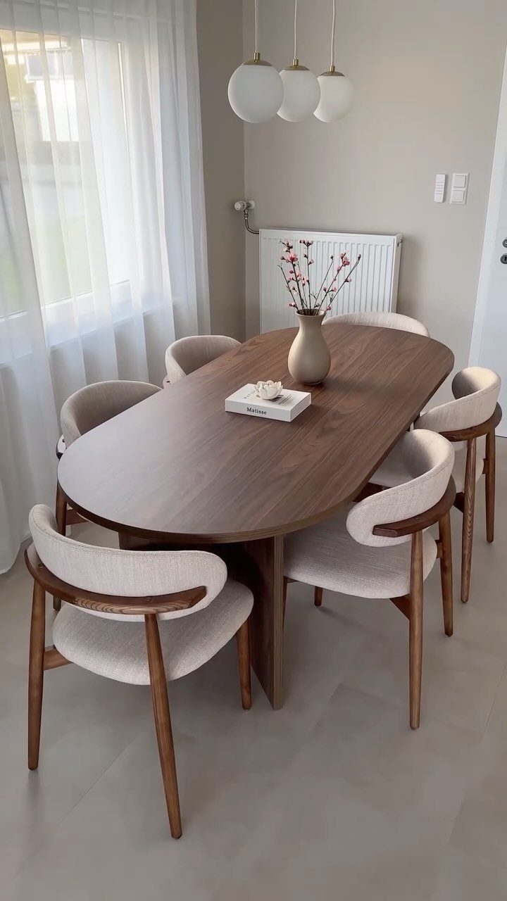 Oval brown wooden dining table with six cream upholstered chairs in a bright room.