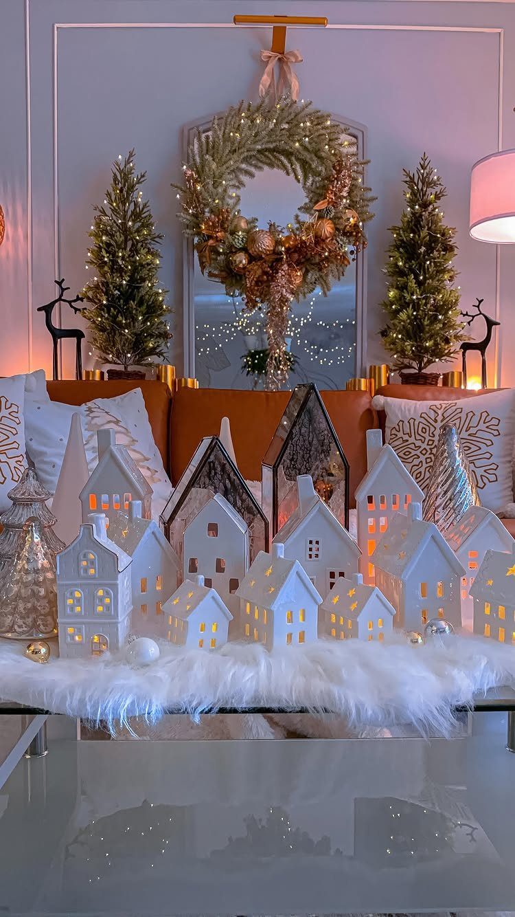 17 Christmas Decor Ideas with White for a Serene Home 6 White ceramic Christmas village with glowing windows and a gold wreath in background.