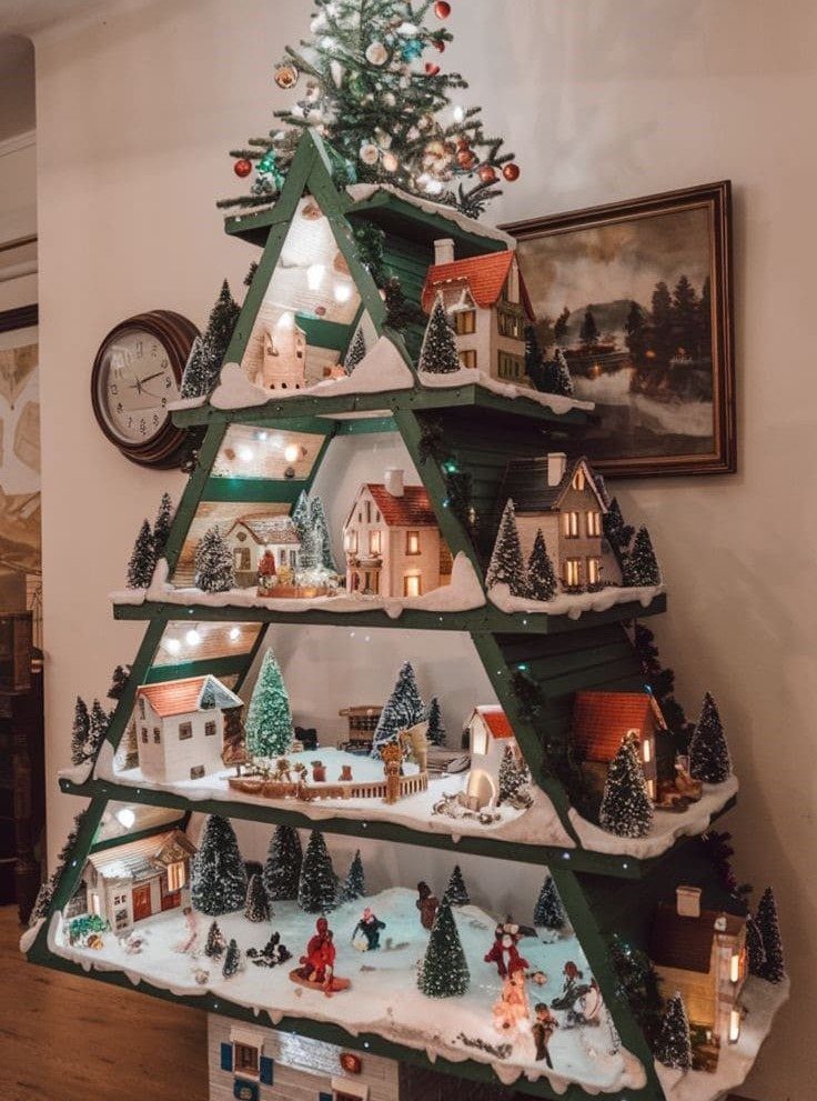 20 Christmas Decorations to Make at Home in 2026 2 Green wooden Christmas tree shelf decorated with miniature snowy village houses and fairy lights.