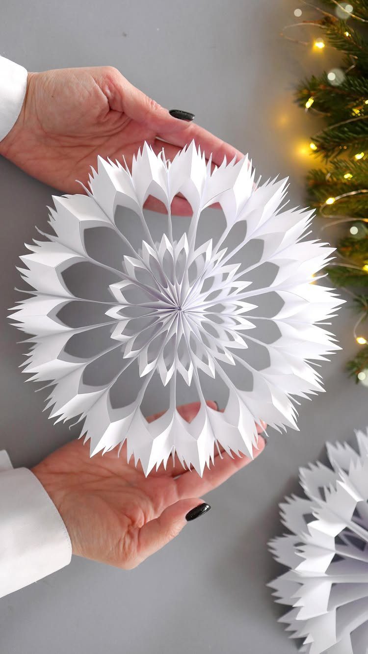 33 Christmas Party Decor Ideas to Bring the Holiday Magic 29 White 3D paper snowflake ornament held by hands with a lit Christmas tree nearby.