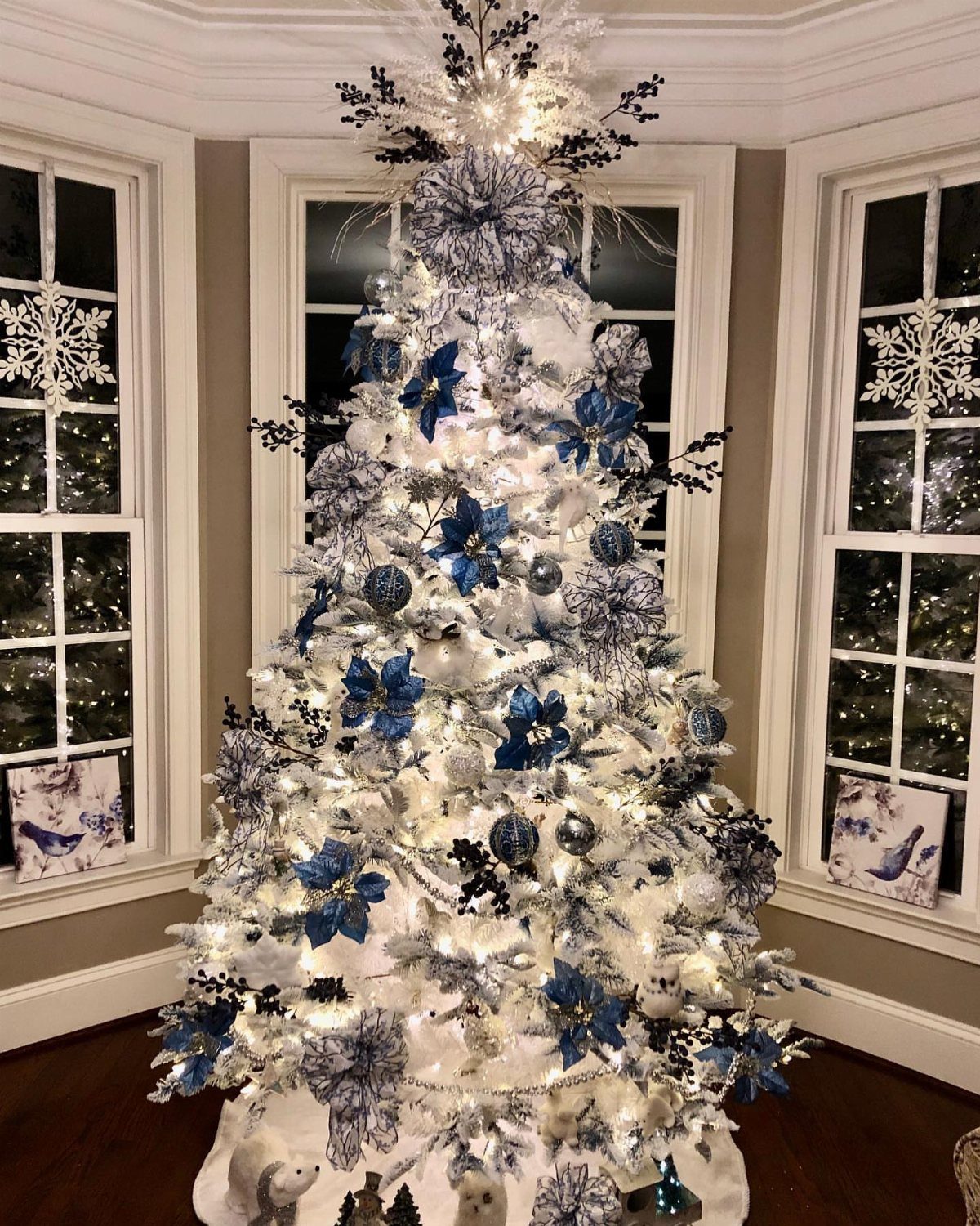 White Christmas tree decorated with blue ornaments and flowers in a bright room.