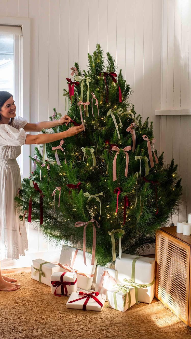 11 Christmas Tree Decorations with Ribbon Ideas 6 Woman attaching a ribbon to a Christmas tree decorated with red and cream bows.