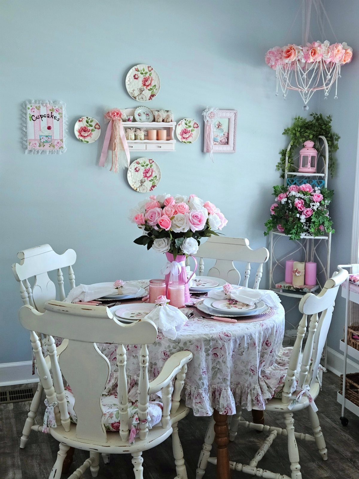 Dining table with floral tablecloth, white chairs, and pink floral decorations in blue room.