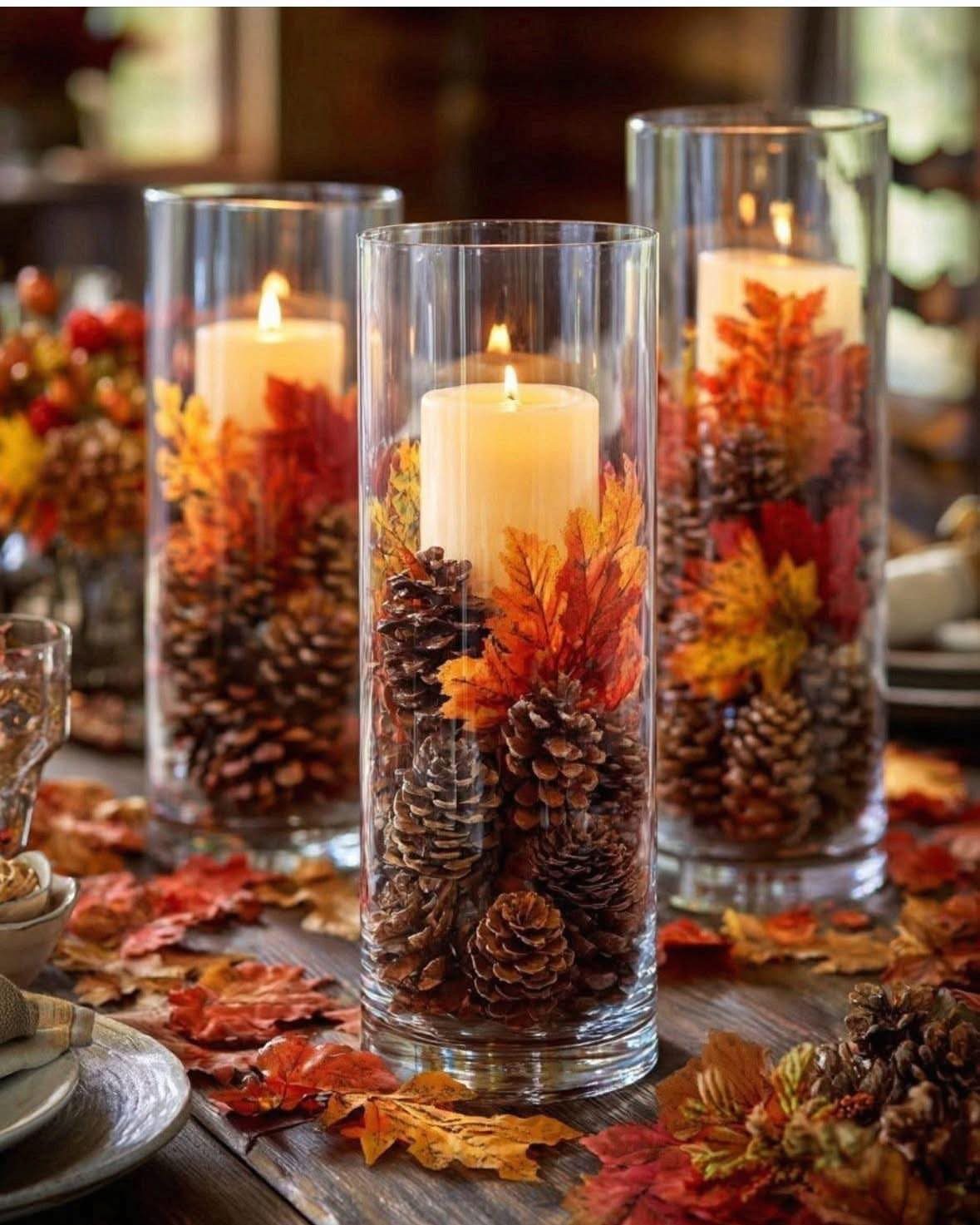 Autumnal candle centerpieces with pinecones and colorful leaves in glass cylinders on wood.