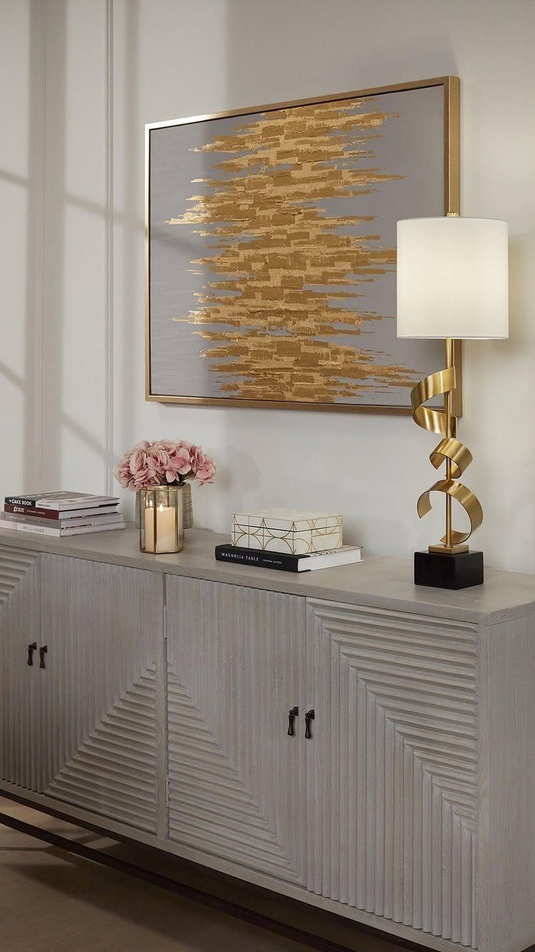 Gold sculptural table lamp on a grey sideboard with abstract art and flowers.