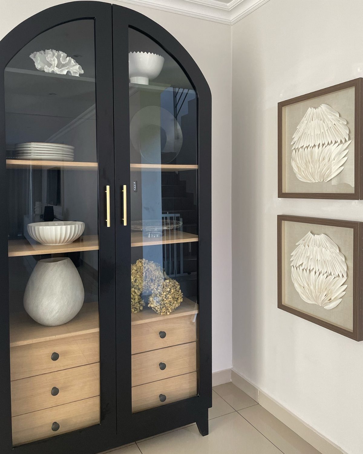 Black arched display cabinet with glass doors and white ceramic decor beside framed art.