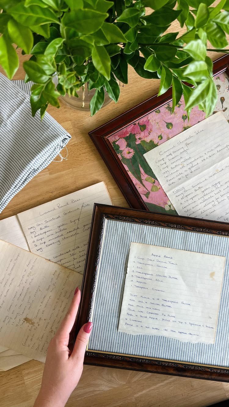 Handwritten recipes in dark wood frames resting on a wooden table with greenery.