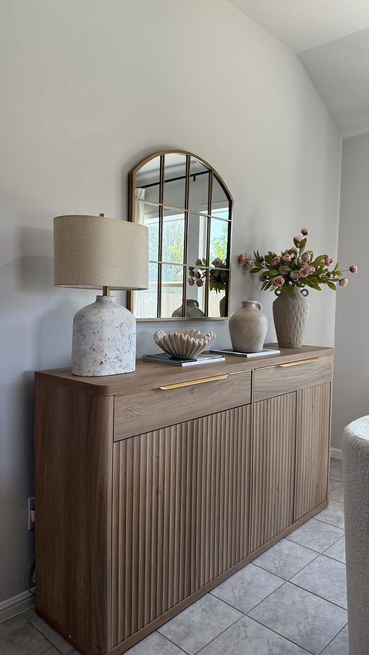 Wooden fluted buffet sideboard with a lamp, vases, and arched mirror above it.