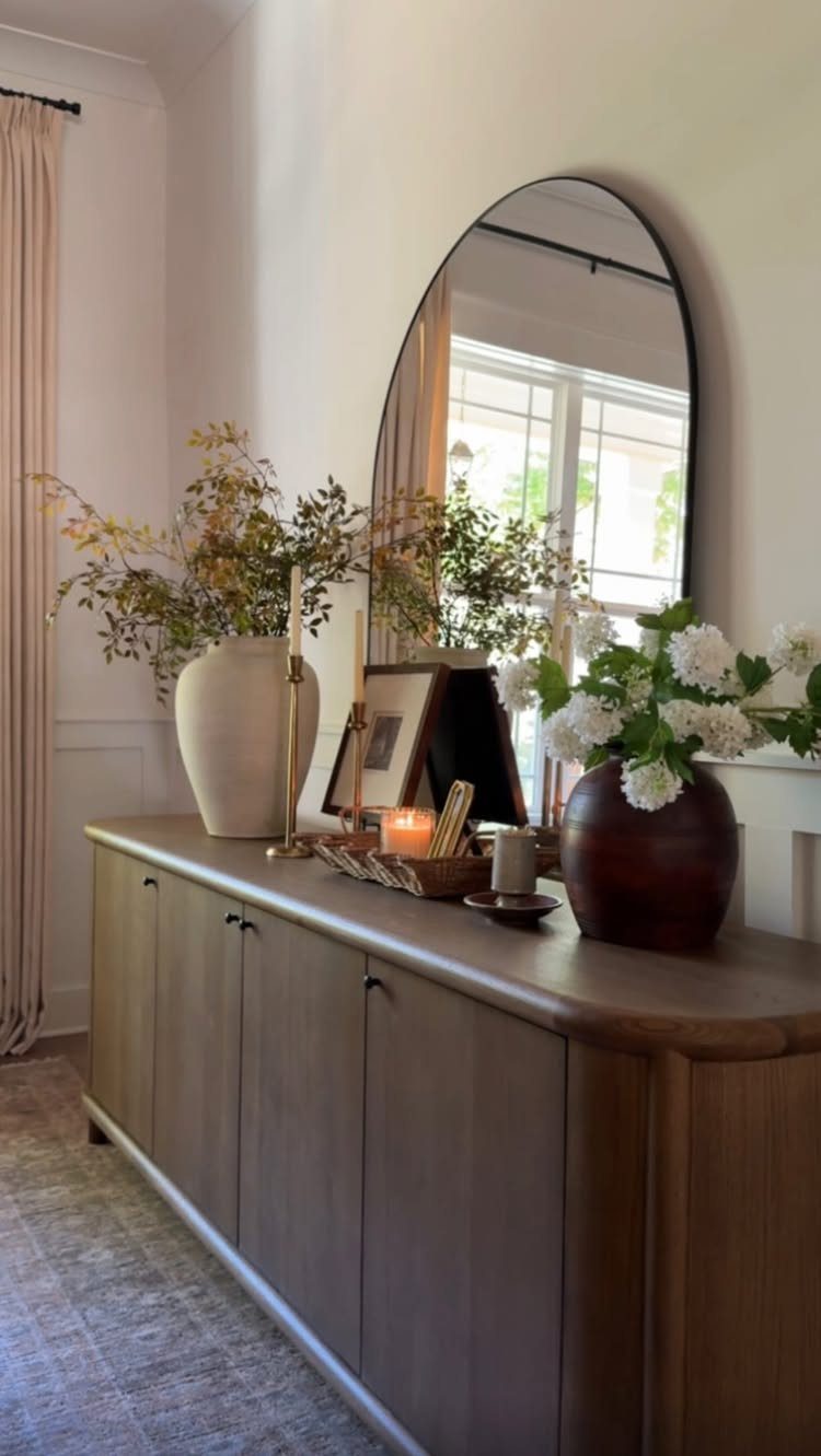 Wooden sideboard with vases, greenery, and candles beneath an arched wall mirror.
