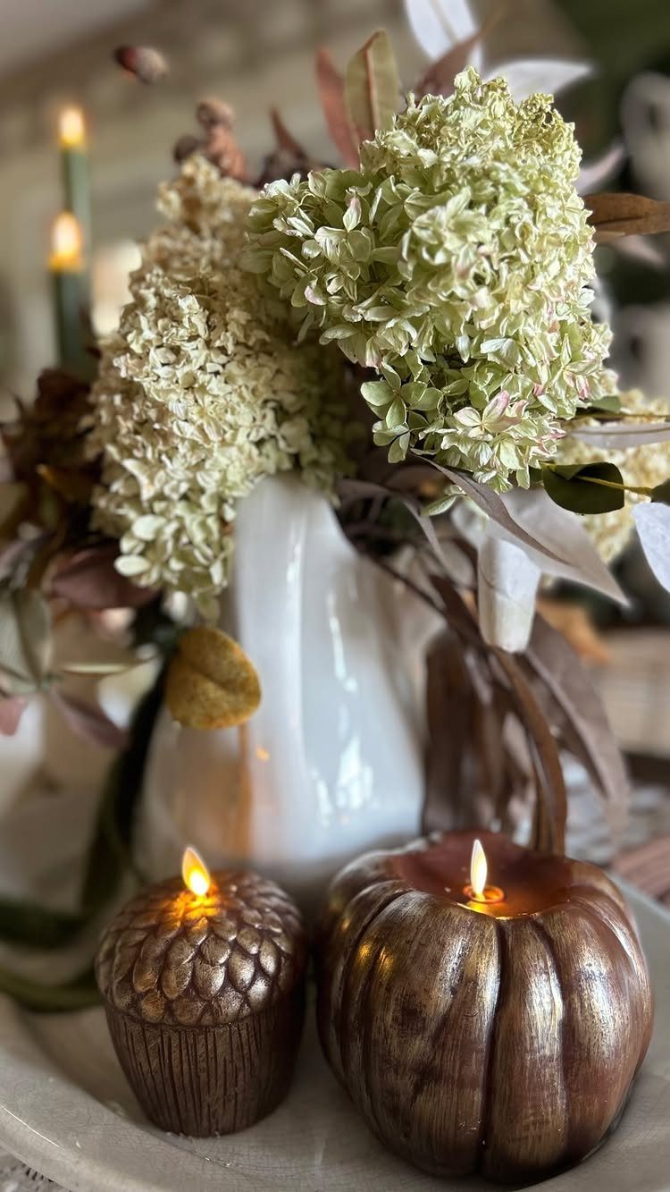Lit pumpkin candles and a white vase with dried hydrangeas on a table.