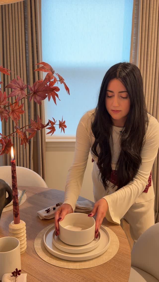 Woman setting a dining table with white dinnerware and autumn leaf decorations.
