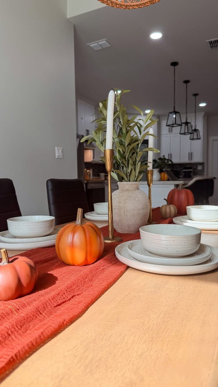 Fall dining table with orange pumpkins, white ceramics, and green foliage in a vase.
