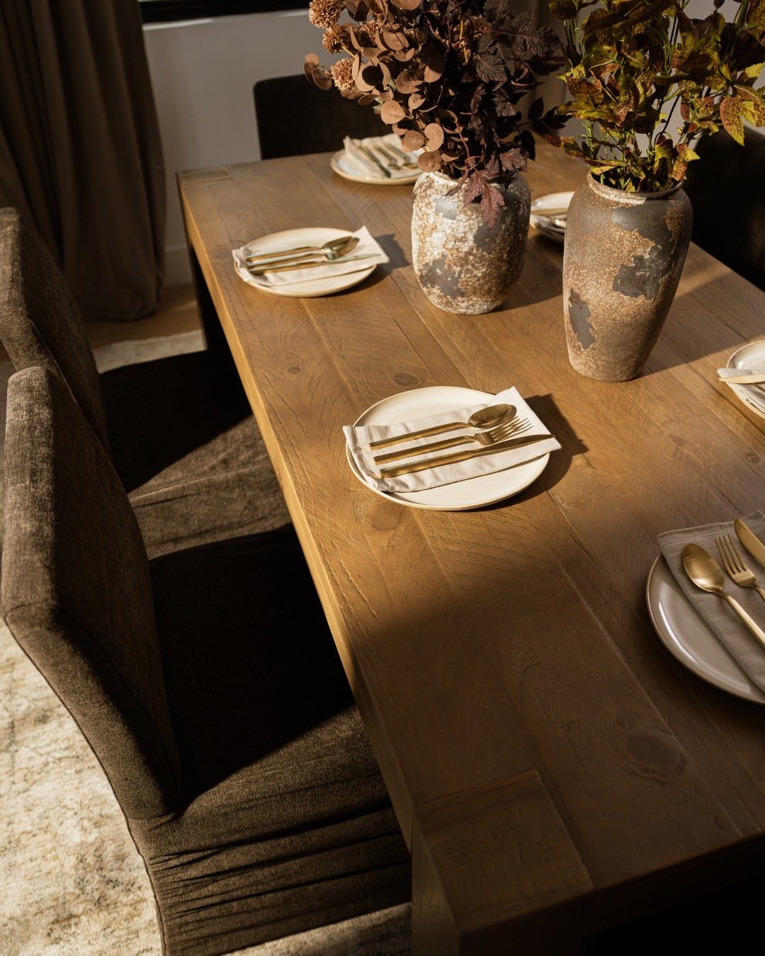 Wooden dining table set with white plates, gold cutlery, and autumn foliage centerpieces.