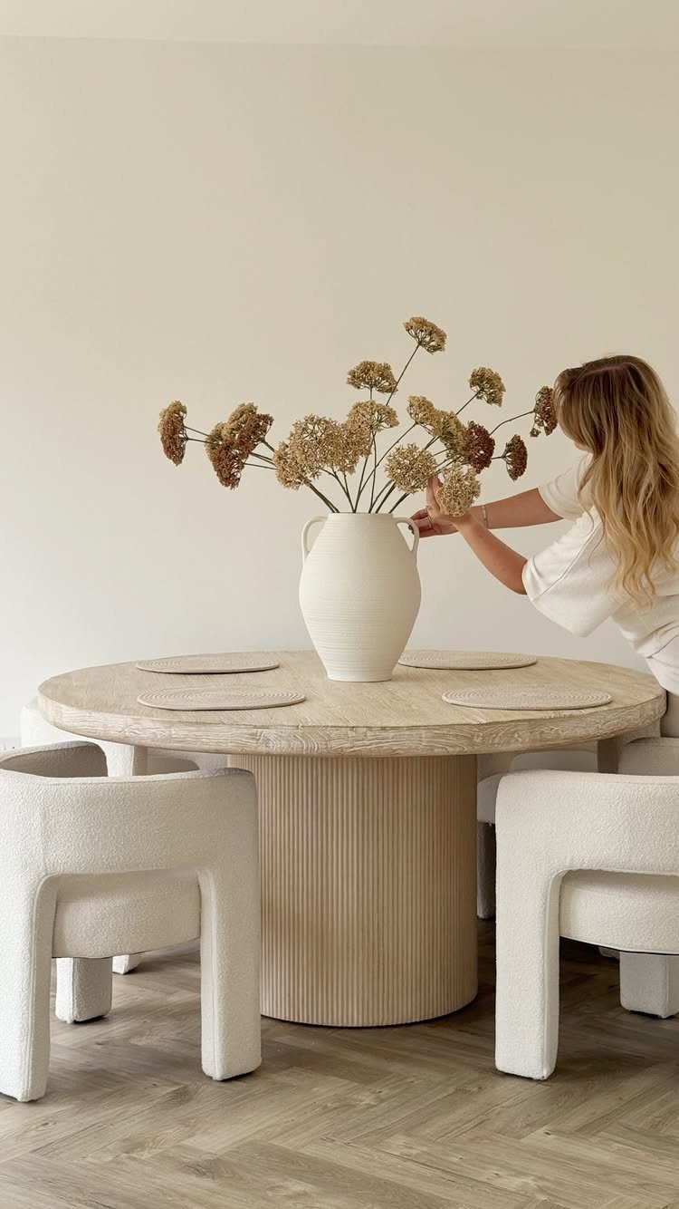 8 Feng Shui Dining Room Ideas for Balance and Flow 2 Woman arranging dried flowers on a round wooden dining table with white chairs.