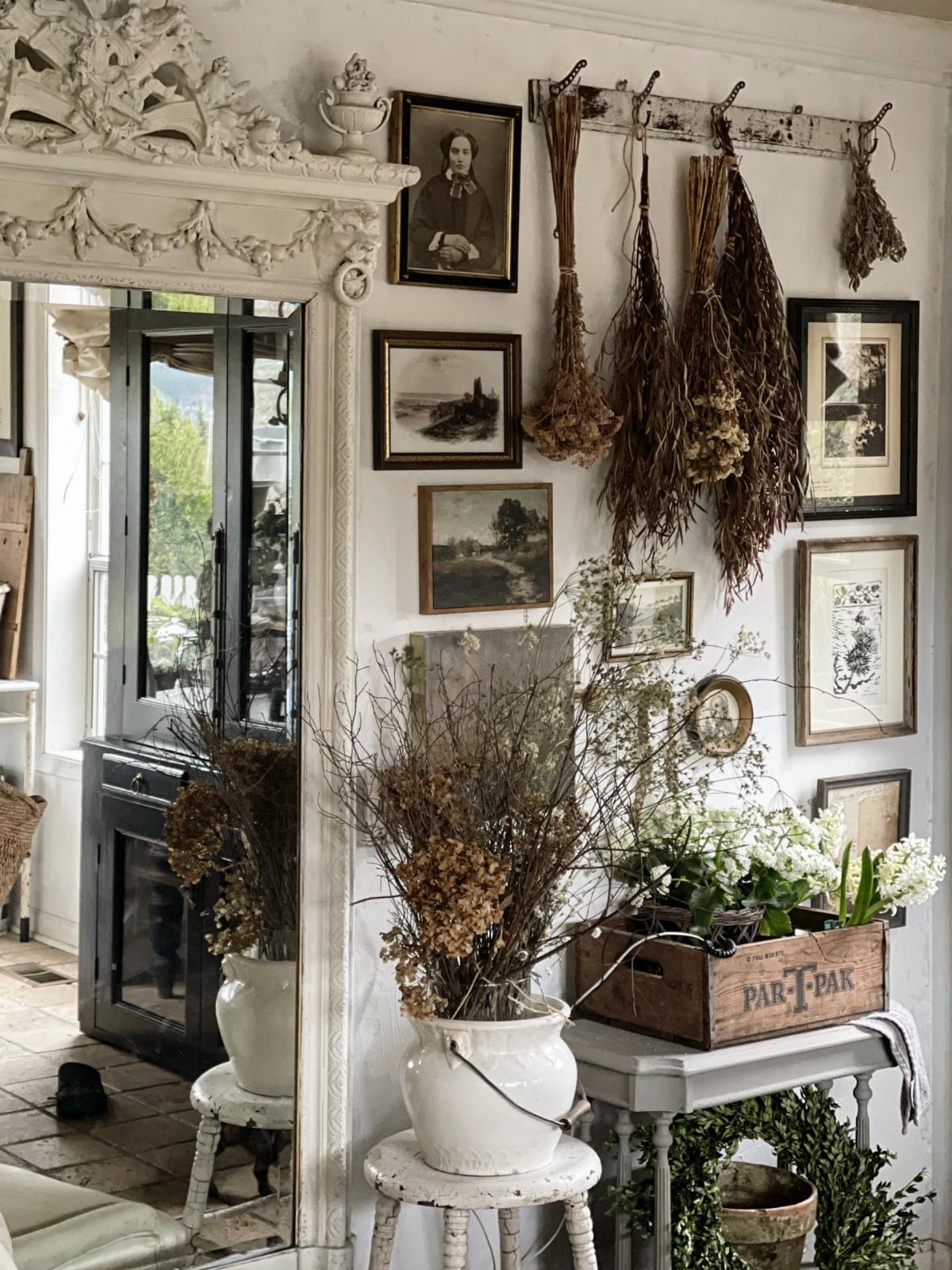 22 French Country Dining Room Ideas with Rustic Charm 21 Framed paintings and portraits on a white wall with hanging dried herbs.