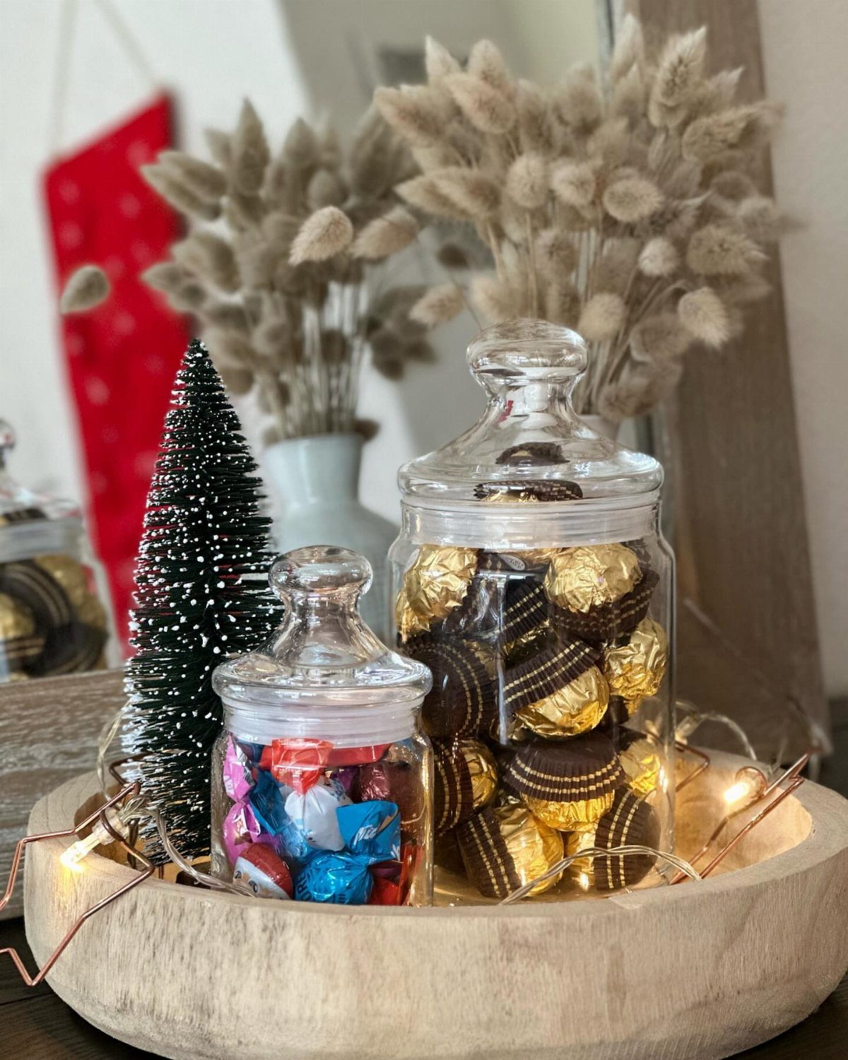 29 New Ideas for Christmas Decorating to Try in 2026 14 Apothecary jars filled with wrapped chocolates in a wooden bowl with fairy lights.