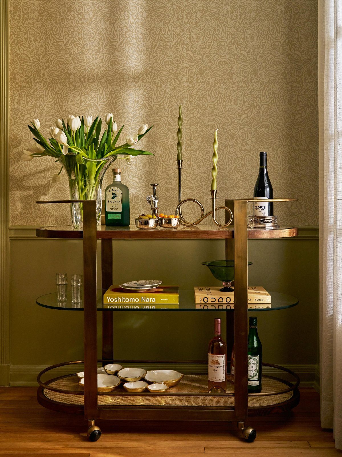 9 Old Money Dining Room Ideas for Timeless Elegance 5 Brass bar cart with wine bottles and white tulips against patterned gold wallpaper.