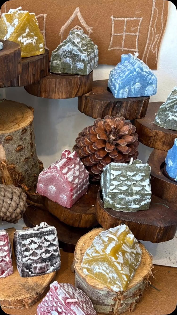 House-shaped holiday candles in various colors displayed on rustic wooden slices with pinecones.