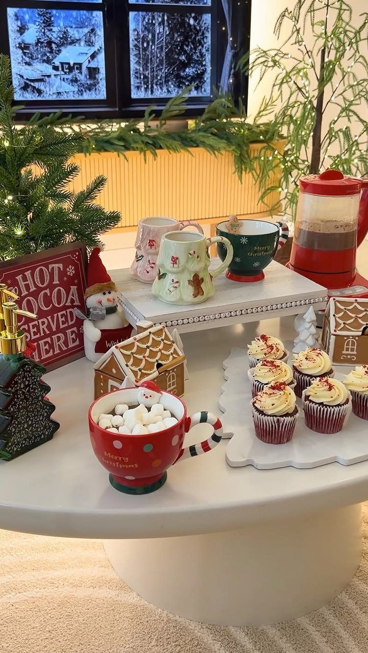 Festive hot chocolate station with red coffee maker, mugs, and gingerbread houses on table.