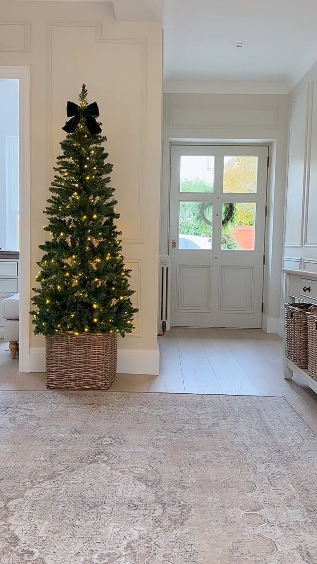 Slender Christmas tree with lights and black bow in a wicker basket hallway.