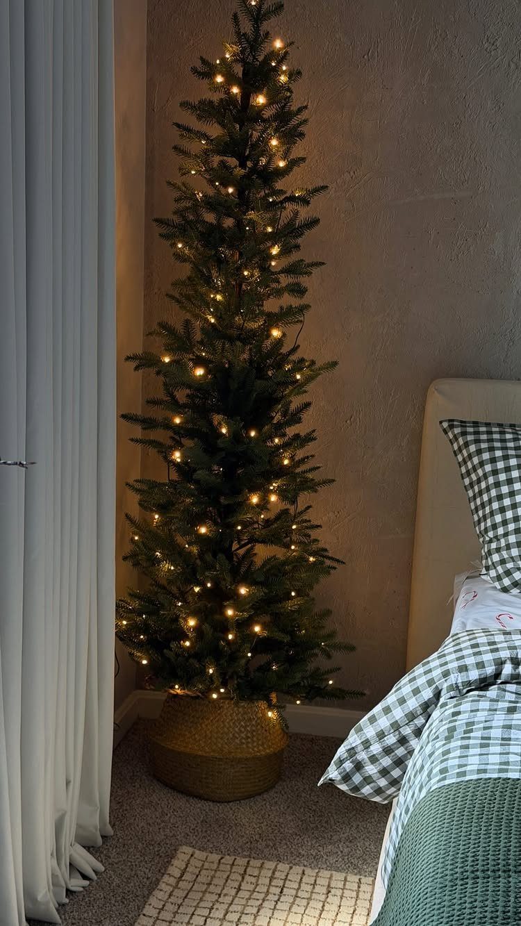 Slim Christmas tree with warm lights in a wicker basket beside a bed.