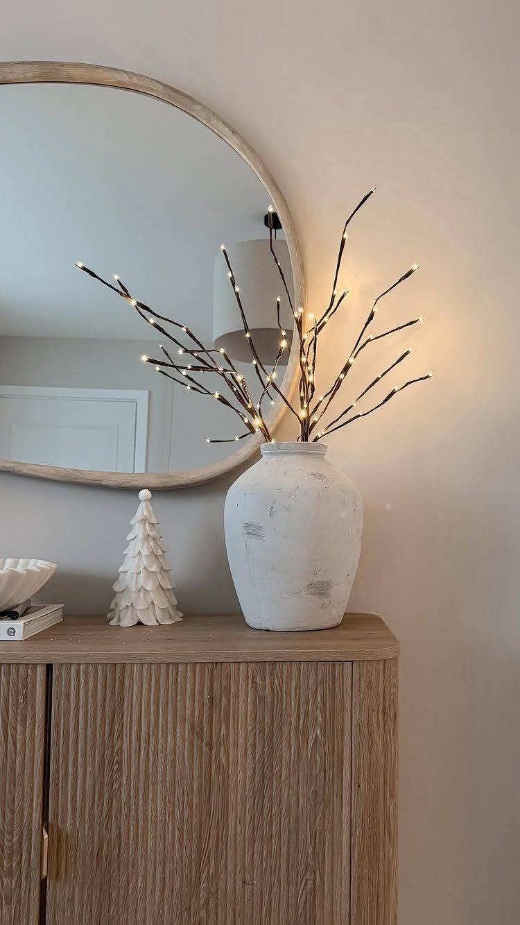 White vase with lit branches and a small white tree on a wooden cabinet.