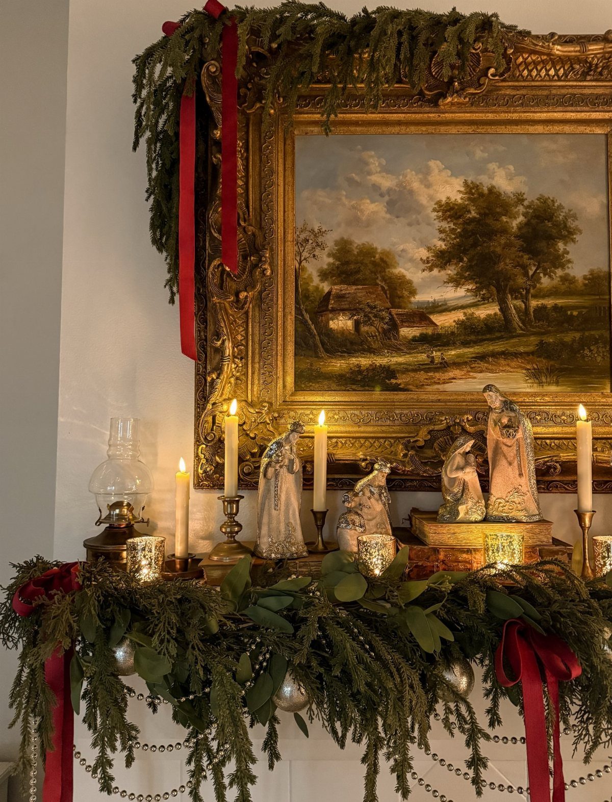 Evergreen garlands with red ribbons decorating a gold-framed painting and a mantelpiece.
