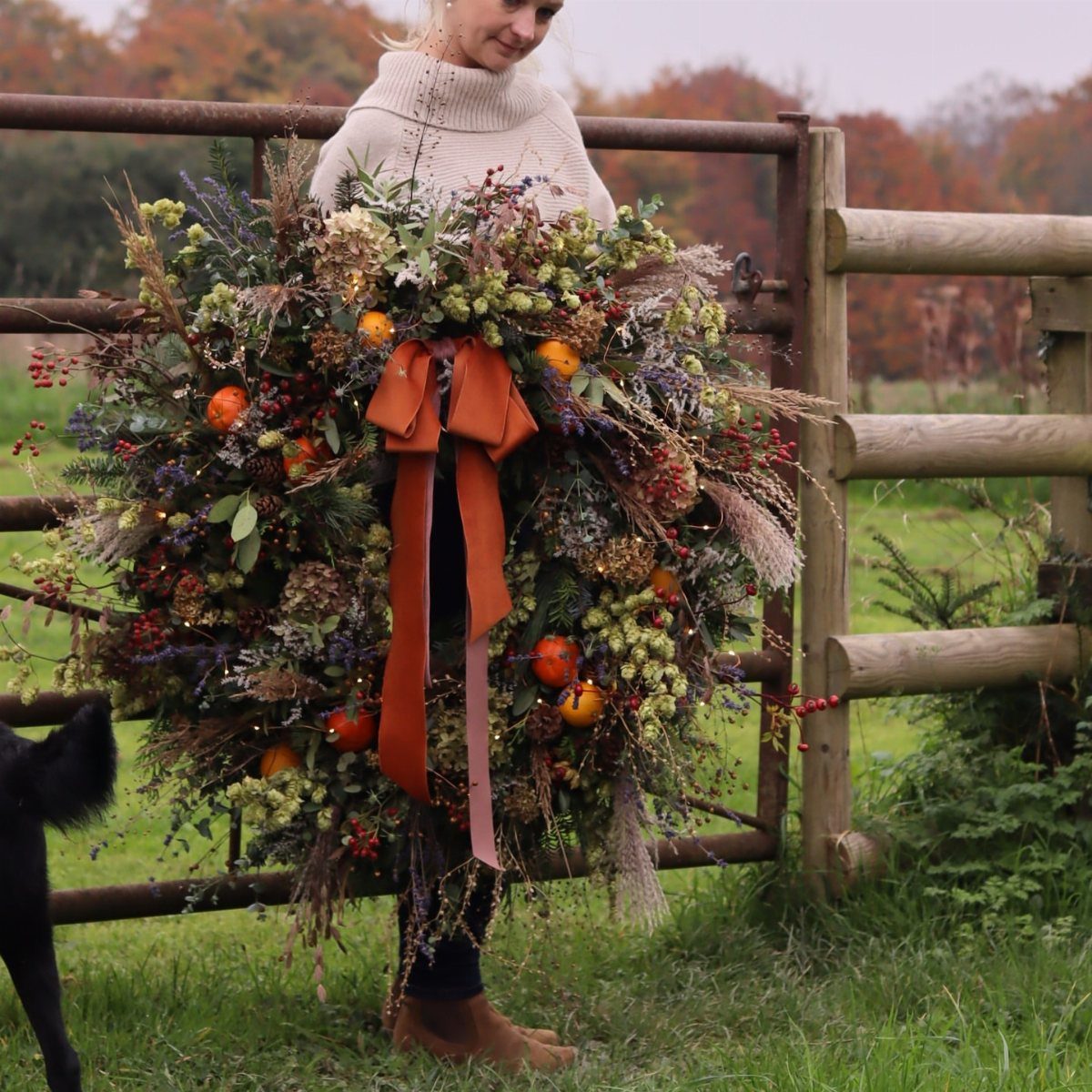 Large natural wreath with greenery, berries, and orange fruits held by a woman.