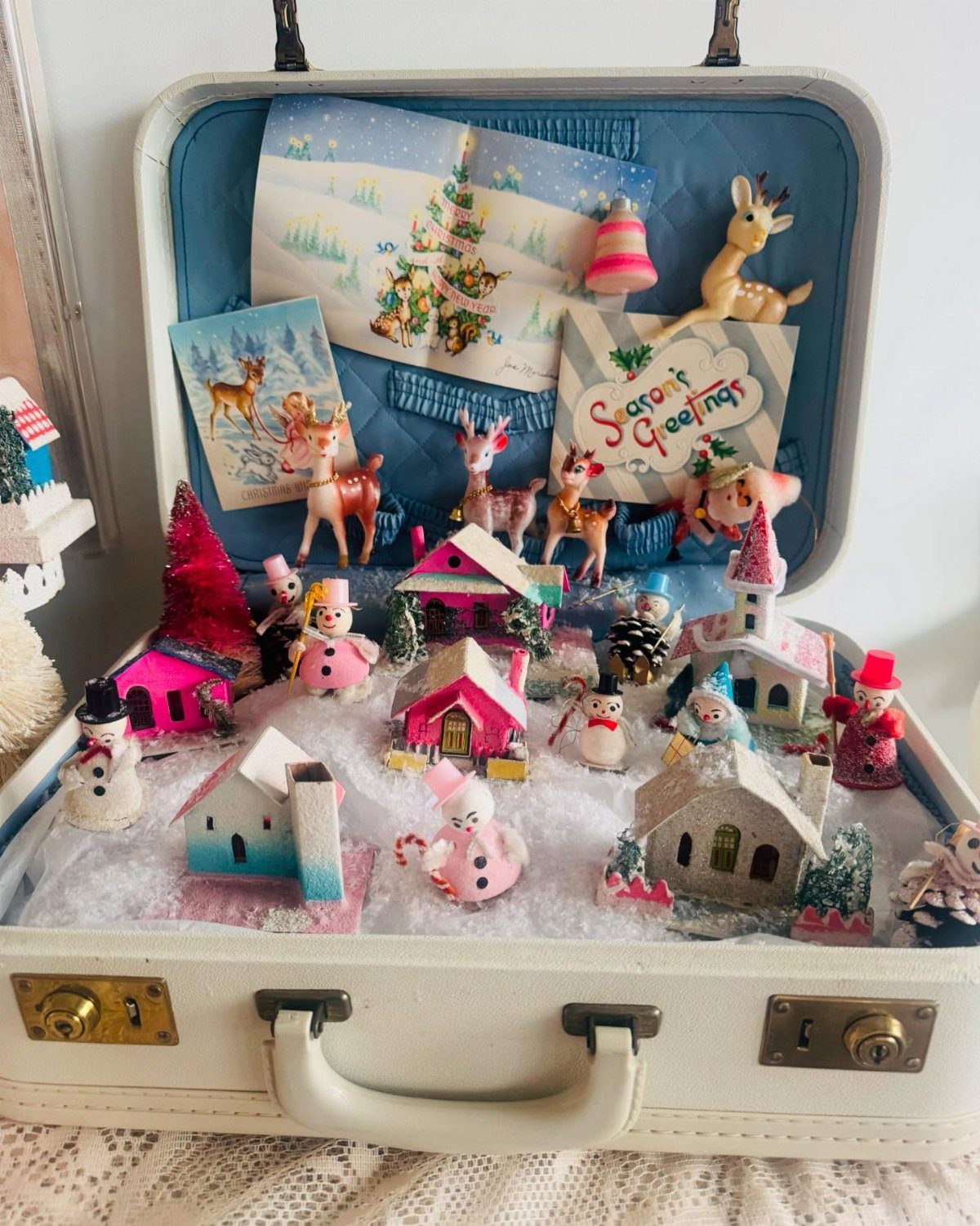 Open white vintage suitcase containing a miniature snowy Christmas village and holiday decorations