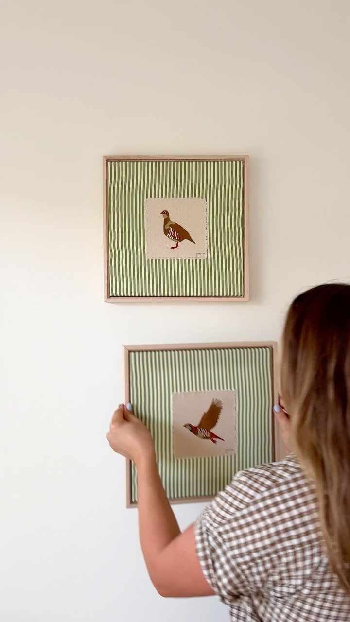 Woman hanging square framed bird prints with green striped mats on white wall