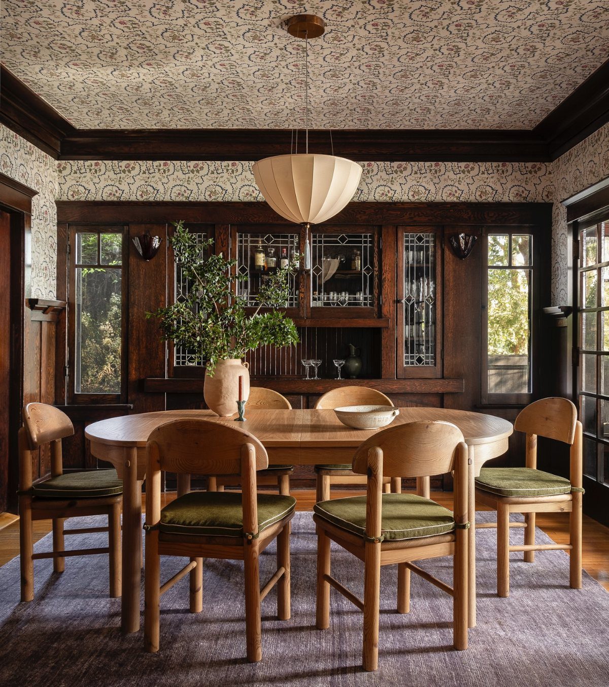 Dining room with dark wood paneling, oval wooden table, and floral wallpaper ceiling.